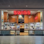 Widok na elegancką witrynę i wejście do salonu z zegarkami marki SWISS w galerii handlowej. Nad wejściem umieszczony jest duży, czerwony, podświetlany napis „SWISS”. W oknach wystawowych oraz na przeszklonych gablotach prezentowane są liczne modele zegarków oraz plakaty reklamowe znanych marek.