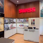 Wnętrze salonu SWISS z widokiem na ladę serwisową oraz przeszklone gabloty ekspozycyjne z zegarkami znanych marek. Na drewnianej ścianie nad ladą zamontowany jest duży, czerwony, podświetlany napis „SWISS”. Na froncie lady serwisowej umieszczono napis „SERWIS PASKÓW” wraz z listą usług: „• Paski do zegarków”, „• Skracanie bransolet”, „• Wymiana pasków” oraz adres strony internetowej „swiss.com.pl butik online”.