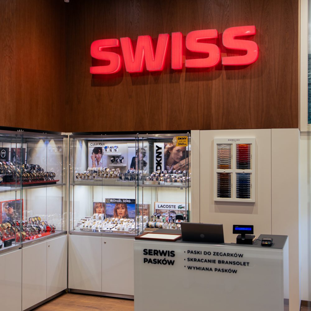 Wnętrze salonu SWISS z widokiem na stanowisko serwisowe i przeszklone gabloty ekspozycyjne z zegarkami znanych marek (m.in. Calvin Klein, DKNY, Michael Kors, Lacoste). Nad ladą serwisową widnieje duży, czerwony, podświetlany napis „SWISS” zamontowany na drewnianej ścianie. Na froncie lady umieszczono napis „SERWIS PASKÓW” oraz informację o usługach: „• Paski do zegarków”, „• Skracanie bransolet”, „• Wymiana pasków”.