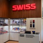 Wnętrze salonu SWISS z widokiem na stanowisko serwisowe i przeszklone gabloty ekspozycyjne z zegarkami znanych marek (m.in. Calvin Klein, DKNY, Michael Kors, Lacoste). Nad ladą serwisową widnieje duży, czerwony, podświetlany napis „SWISS” zamontowany na drewnianej ścianie. Na froncie lady umieszczono napis „SERWIS PASKÓW” oraz informację o usługach: „• Paski do zegarków”, „• Skracanie bransolet”, „• Wymiana pasków”.