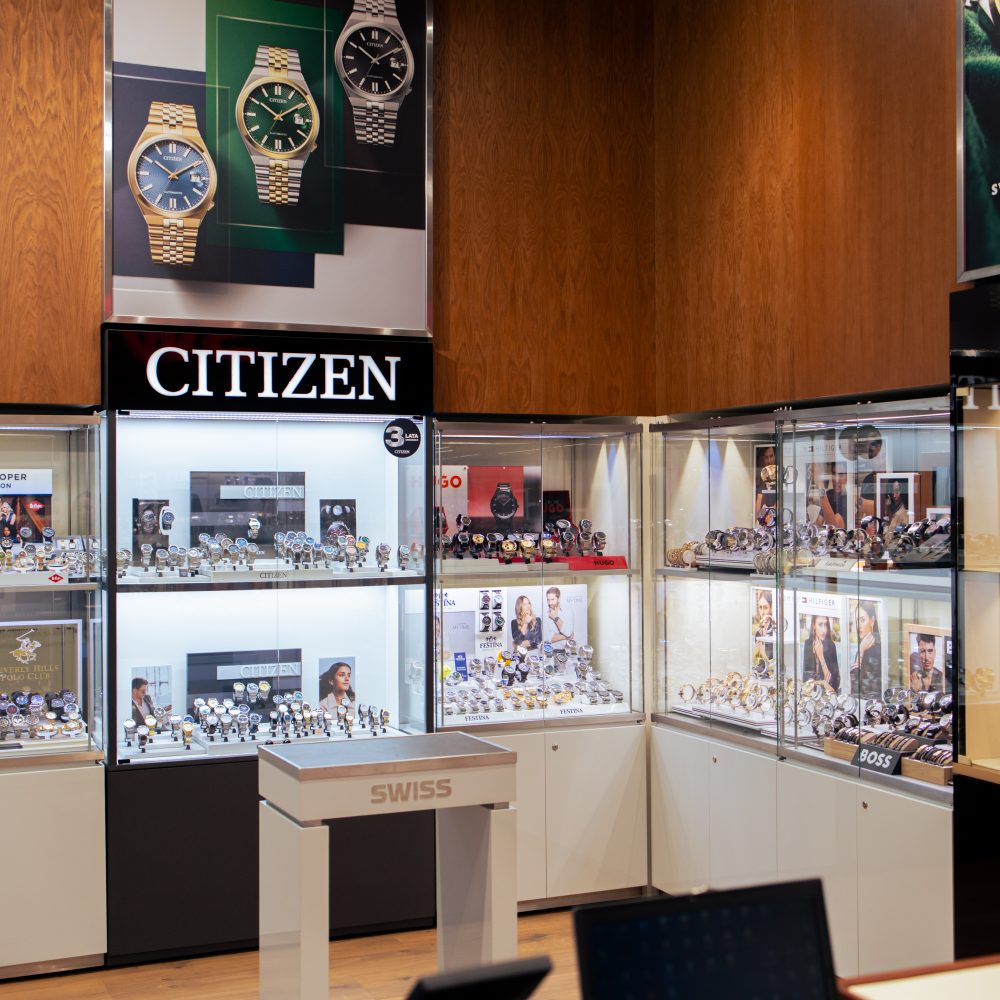 Fragment wnętrza salonu SWISS z dedykowaną ekspozycją zegarków marki Citizen. Nad gablotami znajduje się podświetlany napis „CITIZEN” oraz plakat reklamowy prezentujący złote i srebrne modele zegarków z kolorowymi tarczami. W gablotach i na półkach umieszczono liczne zegarki różnych marek, a na pierwszym planie widoczny jest mały stolik ekspozycyjny z logo SWISS.
