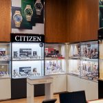 Fragment wnętrza salonu SWISS z dedykowaną ekspozycją zegarków marki Citizen. Nad gablotami znajduje się podświetlany napis „CITIZEN” oraz plakat reklamowy prezentujący złote i srebrne modele zegarków z kolorowymi tarczami. W gablotach i na półkach umieszczono liczne zegarki różnych marek, a na pierwszym planie widoczny jest mały stolik ekspozycyjny z logo SWISS.