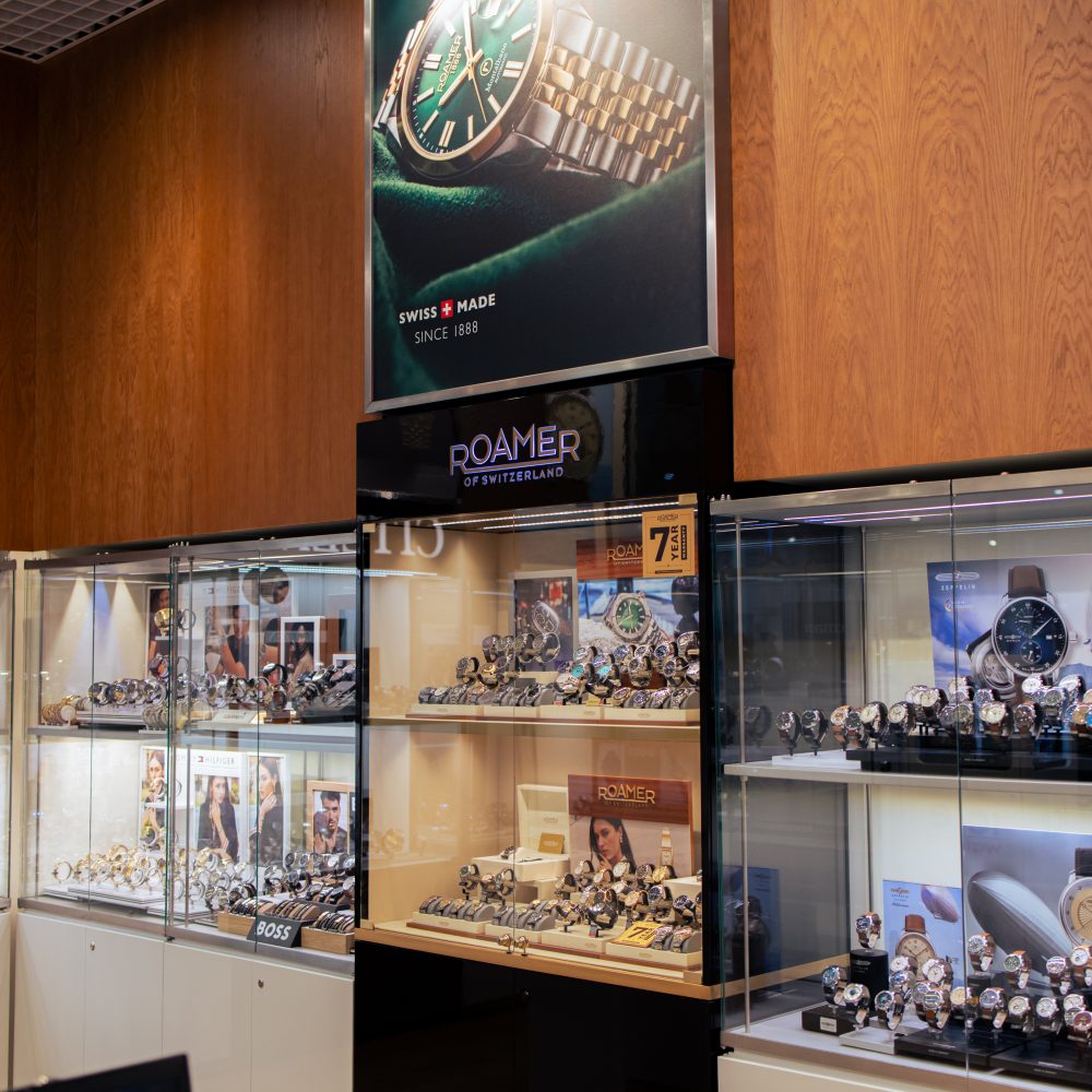 Fragment wnętrza salonu SWISS z ekspozycją szwajcarskich zegarków marki Roamer. Nad gablotami zamontowano duży plakat reklamowy przedstawiający model zegarka z zieloną tarczą oraz napis „SWISS MADE SINCE 1888”. W przeszklonych witrynach wyeksponowano liczne modele zegarków na stojakach oraz firmowe materiały promocyjne marki Roamer.