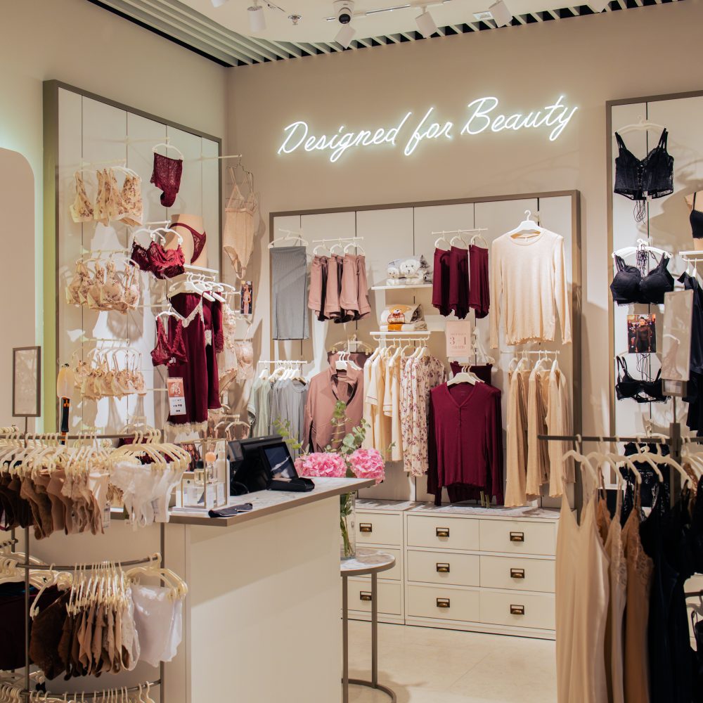 Wnętrze salonu Intimisimi z elegancką ekspozycją damskiej bielizny i odzieży domowej. Nad regałami z produktami widnieje podświetlony na biało napis „Designed for Beauty”. W centralnej części znajduje się jasna lada kasowa, a wokół niej na wieszakach i półkach wyeksponowane są kolekcje bielizny w odcieniach beżu, burgundu i czerni.