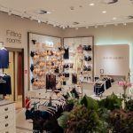 Wnętrze salonu Intimissimi z widocznym wejściem do przymierzalni („Fitting rooms”) oznaczonym po lewej stronie. W centralnej części i na regałach wyeksponowana jest szeroka gama damskiej bielizny w odcieniach beżu, bieli i czerni, a na ścianie widnieje logo marki.