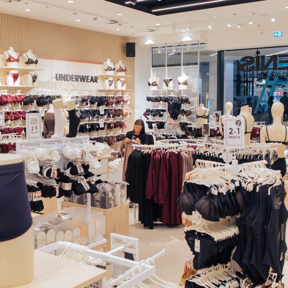 Wnętrze salonu Tezenis z bogatą ekspozycją damskiej bielizny na wieszakach i półkach. Na ścianach i stojakach widoczne są tabliczki informujące o promocji „2+1 gratis” na biustonosze. W głębi sklepu stoi klientka przeglądająca asortyment, a wokół rozmieszczone są manekiny prezentujące kolekcje bielizny.