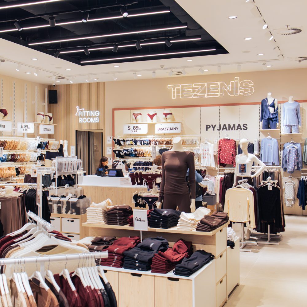 Wnętrze salonu Tezenis z widocznym podświetlanym logo marki na ścianie oraz sekcjami takimi jak „Fitting rooms” i „Pyjamas”. Na stojakach i stołach wyeksponowana jest odzież domowa oraz bielizna w stonowanych kolorach.