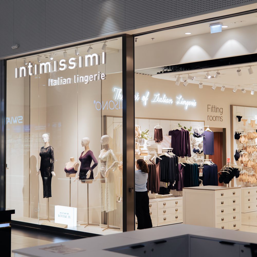 Widok na elegancką witrynę i jasne wnętrze salonu marki Intimissimi w galerii handlowej. W oknie wystawowym stoją manekiny prezentujące damską odzież w odcieniach czerni, fioletu i jasnego beżu, a nad wejściem widnieje podświetlany napis „Intimissimi Italian Lingerie”. Wnętrze sklepu wyposażone jest w półki i stojaki z kolekcją bielizny oraz lady ekspozycyjne.