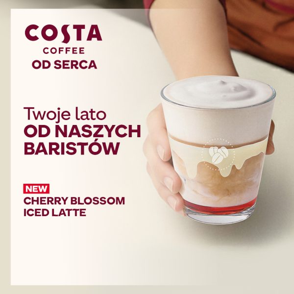 Grafika promocyjna przedstawia dłoń trzymającą szklankę z warstwową kawą mrożoną, która ma na dnie czerwony syrop i gęstą, jasną piankę na górze. Po lewej stronie umieszczono logo Costa Coffee z hasłem „Od serca”, napis „Twoje lato OD NASZYCH BARISTÓW” oraz informację o nowości: „Cherry Blossom Iced Latte”.