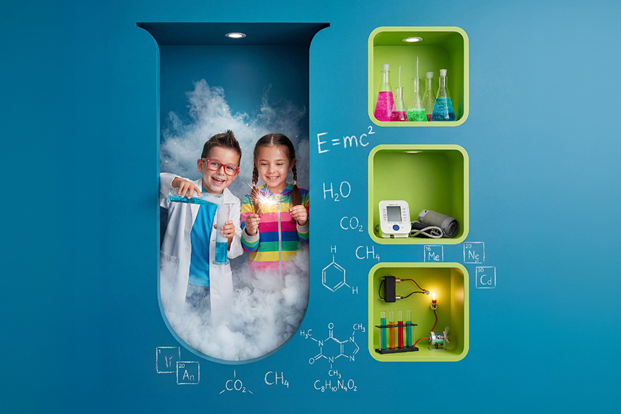 Grafika przedstawia dwoje uśmiechniętych dzieci bawiących się w naukowców wewnątrz wnęki o kształcie kolby laboratoryjnej, wypełnionej efektowną białą parą. Chłopiec w białym fartuchu przelewa niebieski płyn, a dziewczynka trzyma świecącą iskierkę, co nadaje całości magicznego charakteru. Obok znajdują się zielone półki z naczyniami chemicznymi i ciśnieniomierzem, a niebieska ściana ozdobiona jest białymi wzorami chemicznymi i fizycznymi.