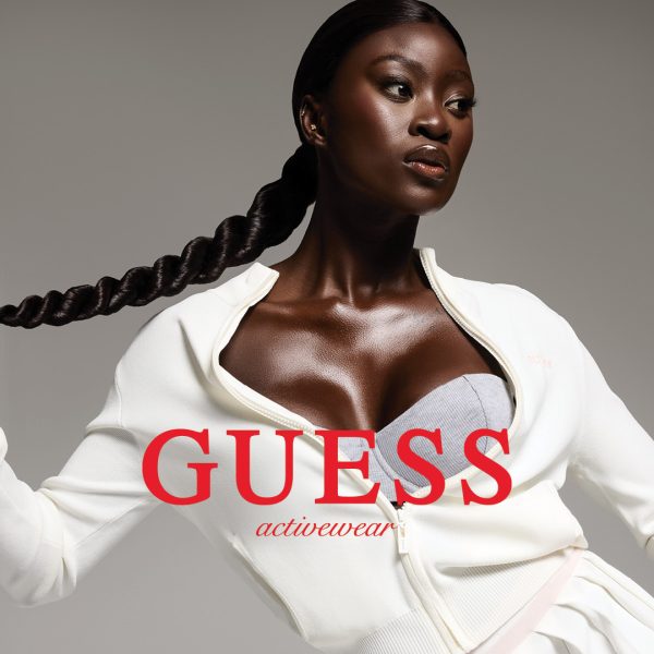 Zdjęcie kampanii Guess Activewear przedstawiające kobietę w białej sportowej bluzie i szarym topie na neutralnym tle. Centralnym elementem jest duży, czerwony logotyp marki „GUESS activewear” nałożony na postać.