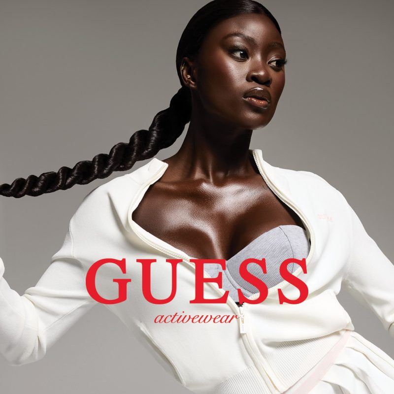 Zdjęcie kampanii Guess Activewear przedstawiające kobietę w białej sportowej bluzie i szarym topie na neutralnym tle. Centralnym elementem jest duży, czerwony logotyp marki „GUESS activewear” nałożony na postać.