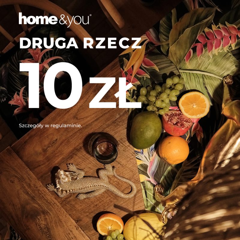 Grafika promocyjna marki home&you informująca o akcji „DRUGA RZECZ 10 ZŁ” na tle nastrojowej aranżacji wnętrza. Zdjęcie przedstawia drewniany stół z tropikalnymi dodatkami, owocami takimi jak mango, winogrona i cytrusy oraz złotą figurką jaszczurki.