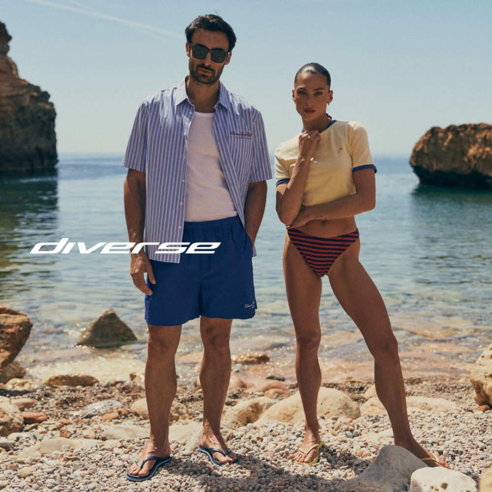 Grafika reklamowa marki Diverse przedstawia kobietę i mężczyznę w letnich stylizacjach plażowych, stojących na kamienistym brzegu morza na tle skalistego krajobrazu. Mężczyzna ma na sobie rozpiętą koszulę w paski i niebieskie szorty, natomiast kobieta pozuje w żółtym t-shircie i pasiaste dół od bikini, a pośrodku kadru umieszczono białe logo marki.