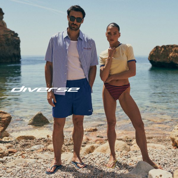 Grafika reklamowa marki Diverse przedstawia kobietę i mężczyznę w letnich stylizacjach plażowych, stojących na kamienistym brzegu morza na tle skalistego krajobrazu. Mężczyzna ma na sobie rozpiętą koszulę w paski i niebieskie szorty, natomiast kobieta pozuje w żółtym t-shircie i pasiaste dół od bikini, a pośrodku kadru umieszczono białe logo marki.