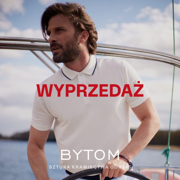Plakat promocyjny marki Bytom przedstawia brodatego mężczyznę w białej koszulce polo, stojącego za sterem jachtu na tle wody i częściowo zachmurzonego nieba. W centralnej części grafiki umieszczono duży, czerwony napis „WYPRZEDAŻ”, a na dole widnieje białe logo marki z hasłem „SZTUKA KRAWIECTWA OD 1945”.