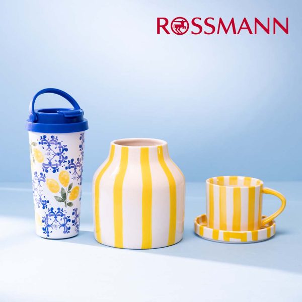 Grafika promocyjna sieci Rossmann przedstawia kolekcję naczyń i akcesoriów marki Eloy, w tym wysoki kubek termiczny z motywem cytryn oraz pękaty wazon i filiżankę ze spodkiem w żółto-białe pasy. Produkty są wyeksponowane na jednolitym, jasnoniebieskim tle, a w prawym górnym rogu umieszczono czerwone logo drogerii.