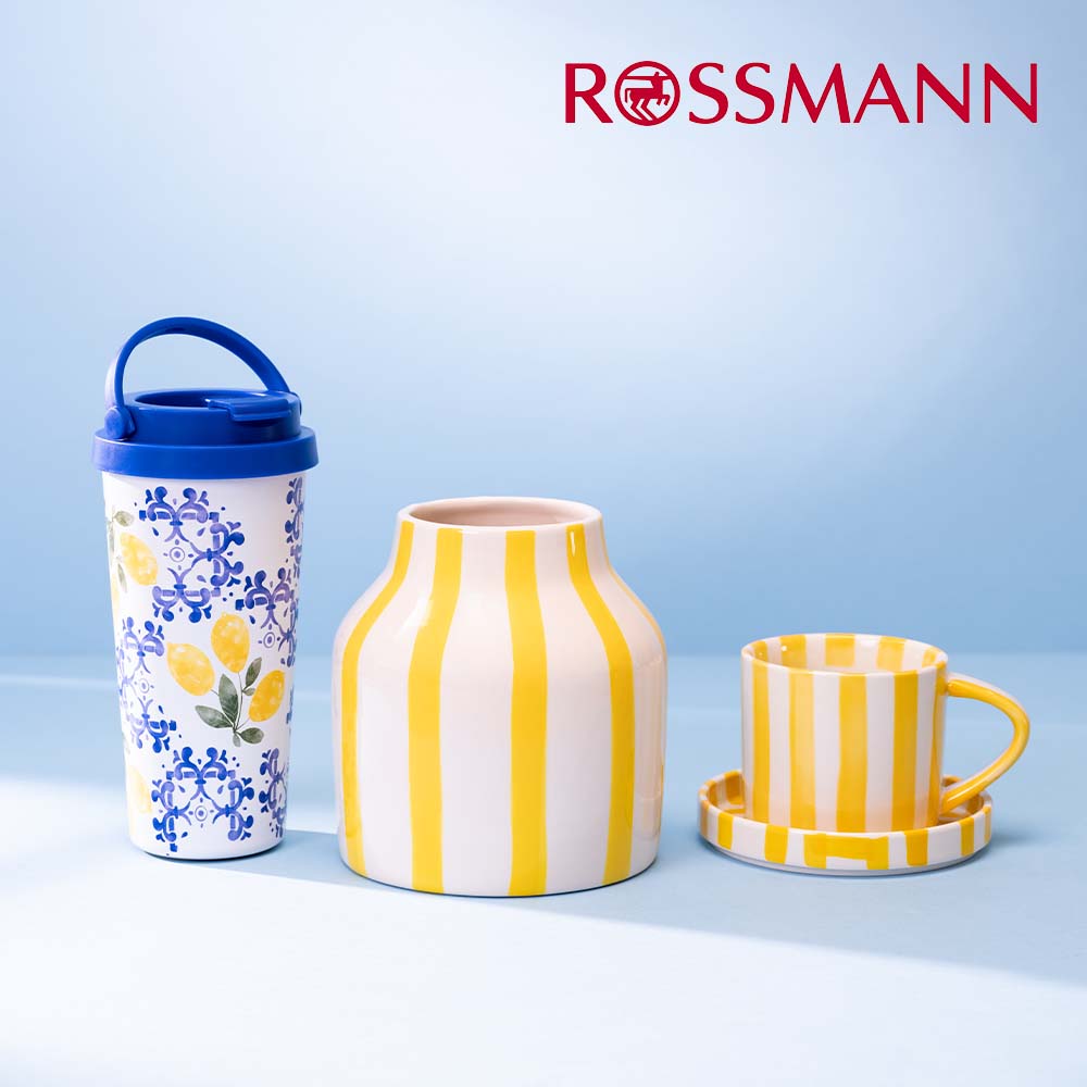 Grafika promocyjna sieci Rossmann przedstawia kolekcję naczyń i akcesoriów marki Eloy, w tym wysoki kubek termiczny z motywem cytryn oraz pękaty wazon i filiżankę ze spodkiem w żółto-białe pasy. Produkty są wyeksponowane na jednolitym, jasnoniebieskim tle, a w prawym górnym rogu umieszczono czerwone logo drogerii.