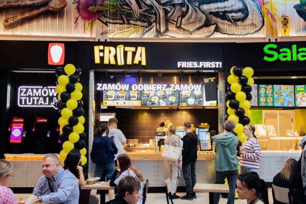 Widok na stoisko gastronomiczne Frita w strefie food court galerii handlowej. Nad ladą umieszczony jest żółto-czarny szyld z nazwą marki „FRITA” oraz hasłem „FRIES.FIRST!”. Wejściowe filtry dekorują girlandy z żółtych i czarnych balonów, a na pierwszym planie widoczni są klienci przy stolikach oraz zamawiający jedzenie przy ladzie.