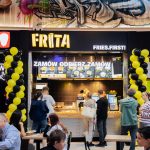 Widok na stoisko gastronomiczne Frita w strefie food court galerii handlowej. Nad ladą umieszczony jest żółto-czarny szyld z nazwą marki „FRITA” oraz hasłem „FRIES.FIRST!”. Wejściowe filtry dekorują girlandy z żółtych i czarnych balonów, a na pierwszym planie widoczni są klienci przy stolikach oraz zamawiający jedzenie przy ladzie.