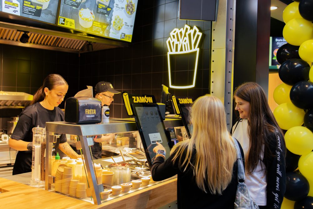 Klienci składający zamówienie przy samoobsługowych ekranach w stoisku gastronomicznym Frita. Nad ladą widoczny jest podświetlany, neonowy szyld w kształcie kubka z frytkami oraz obsługa przygotowująca posiłki w tle.