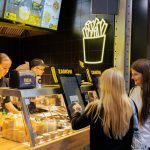 Klienci składający zamówienie przy samoobsługowych ekranach w stoisku gastronomicznym Frita. Nad ladą widoczny jest podświetlany, neonowy szyld w kształcie kubka z frytkami oraz obsługa przygotowująca posiłki w tle.