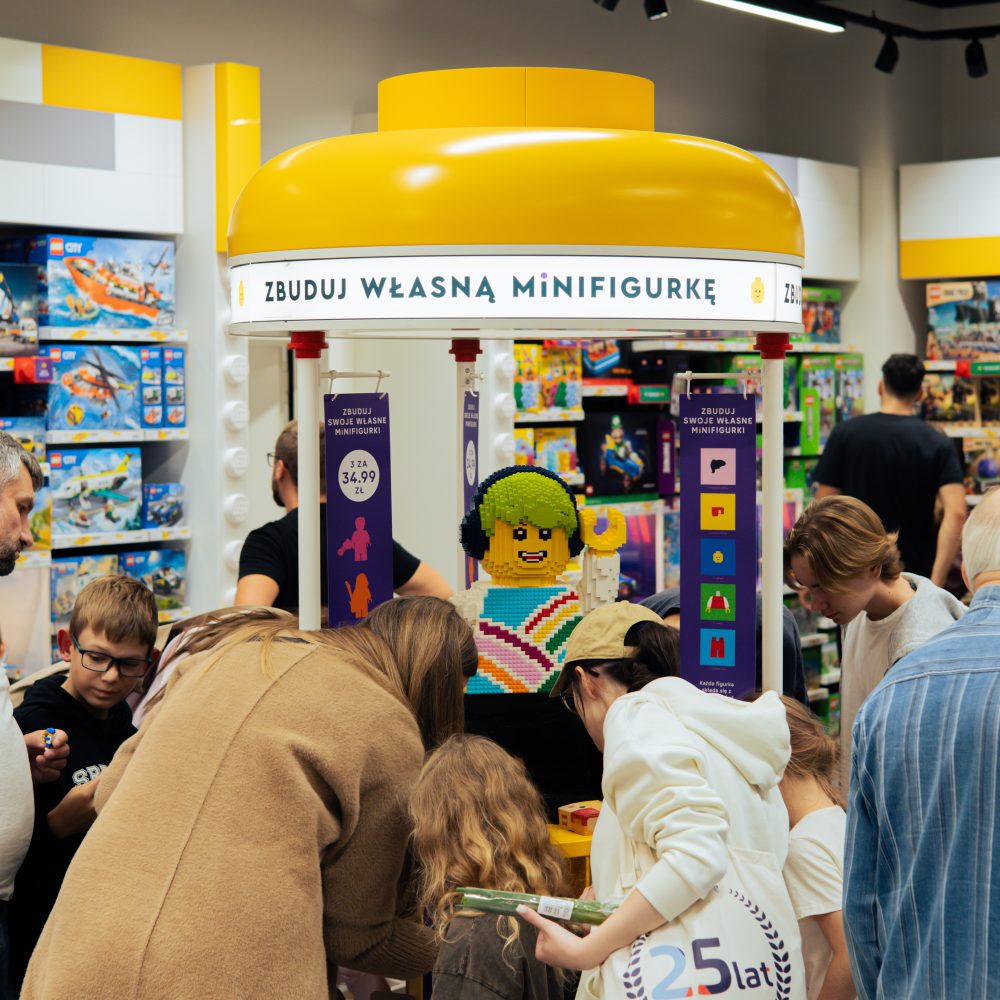 Stanowisko „Zbuduj własną minifigurkę” w salonie LEGO, wokół którego gromadzą się klienci i dzieci. Nad stanowiskiem widoczny jest duży, żółty element w kształcie klocka z podświetlanym napisem informacyjnym oraz wiszące banery promocyjne z ceną zestawu.