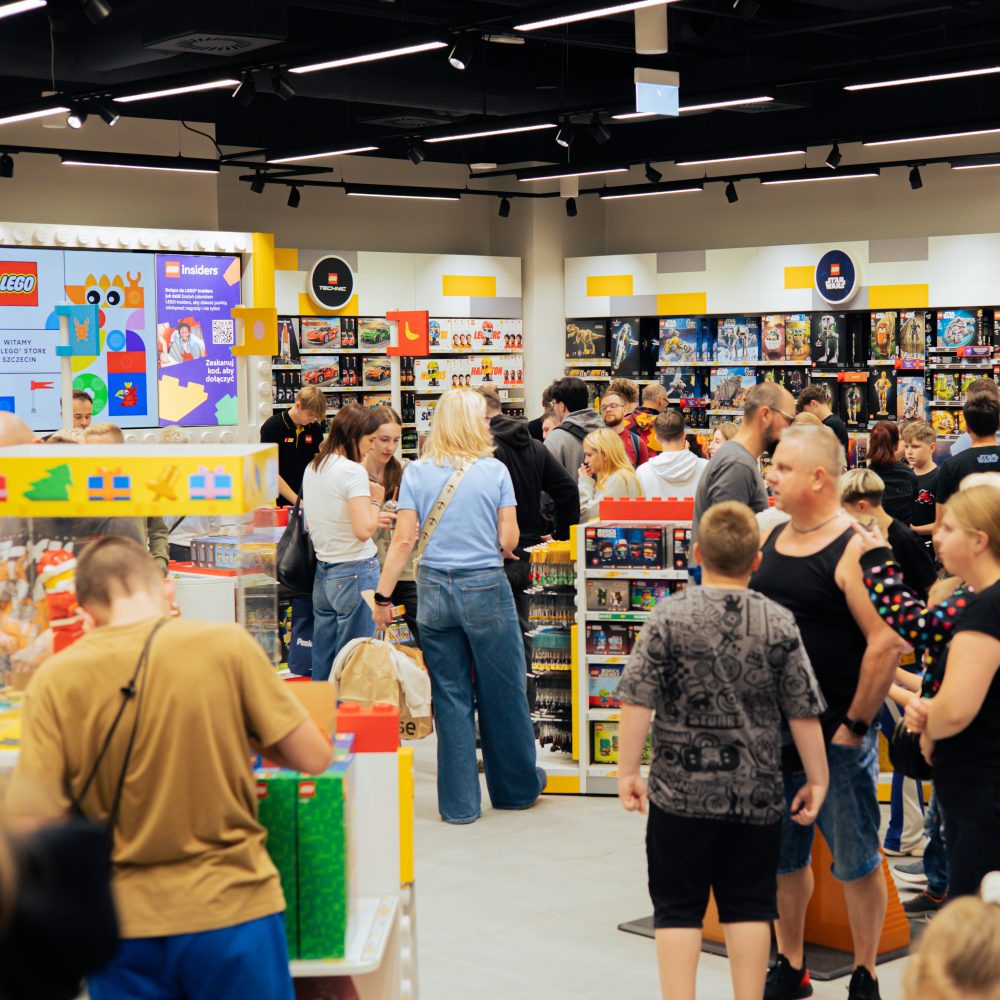 Wnętrze zatłoczonego salonu LEGO z klientami przeglądającymi asortyment zestawów klocków na regałach i ekspozytorach. Na ścianach widoczne są kolorowe panele reklamowe programu LEGO Insiders oraz logotypy poszczególnych serii produktów.