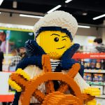 Duża figurka marynarza zbudowana z klocków LEGO, znajdująca się wewnątrz salonu. Postać w białej czapce marynarskiej i granatowym stroju stoi przy brązowym kole sterowym, trzymając je w rękach.
