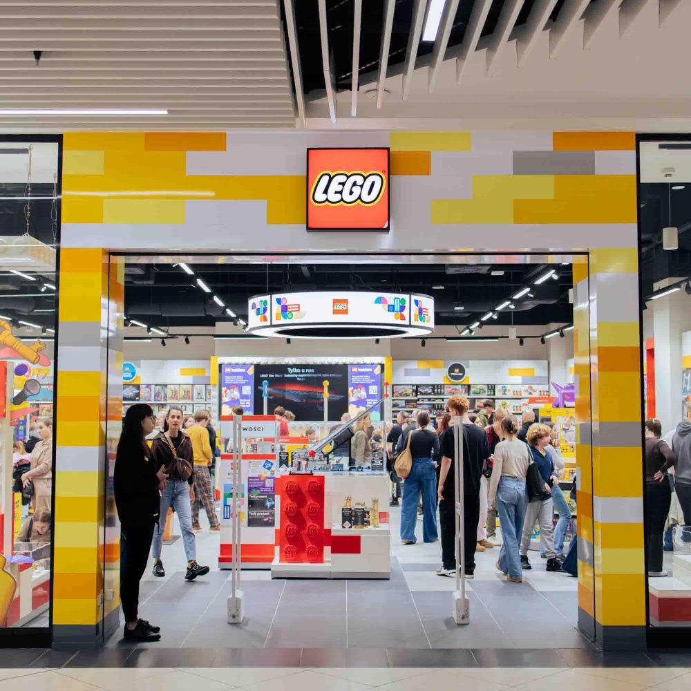 Widok na wejście i ruchliwe wnętrze oficjalnego salonu marki LEGO w galerii handlowej. Fasada sklepu ozdobiona jest charakterystycznym motywem kolorowych, żółtych, biało-szarych i pomarańczowych klocków oraz centralnie umieszczonym, podświetlanym logo LEGO w czerwonej ramce. Wewnątrz widoczne są liczne ekspozycje zestawów, regały z produktami oraz klienci i pracownicy.