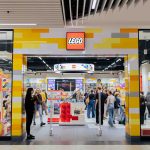 Widok na wejście i ruchliwe wnętrze oficjalnego salonu marki LEGO w galerii handlowej. Fasada sklepu ozdobiona jest charakterystycznym motywem kolorowych, żółtych, biało-szarych i pomarańczowych klocków oraz centralnie umieszczonym, podświetlanym logo LEGO w czerwonej ramce. Wewnątrz widoczne są liczne ekspozycje zestawów, regały z produktami oraz klienci i pracownicy.